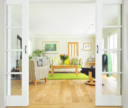 Looking through glas doors into a living room with light wood floors a small green rug, beige chairs and a wooden coffee table.